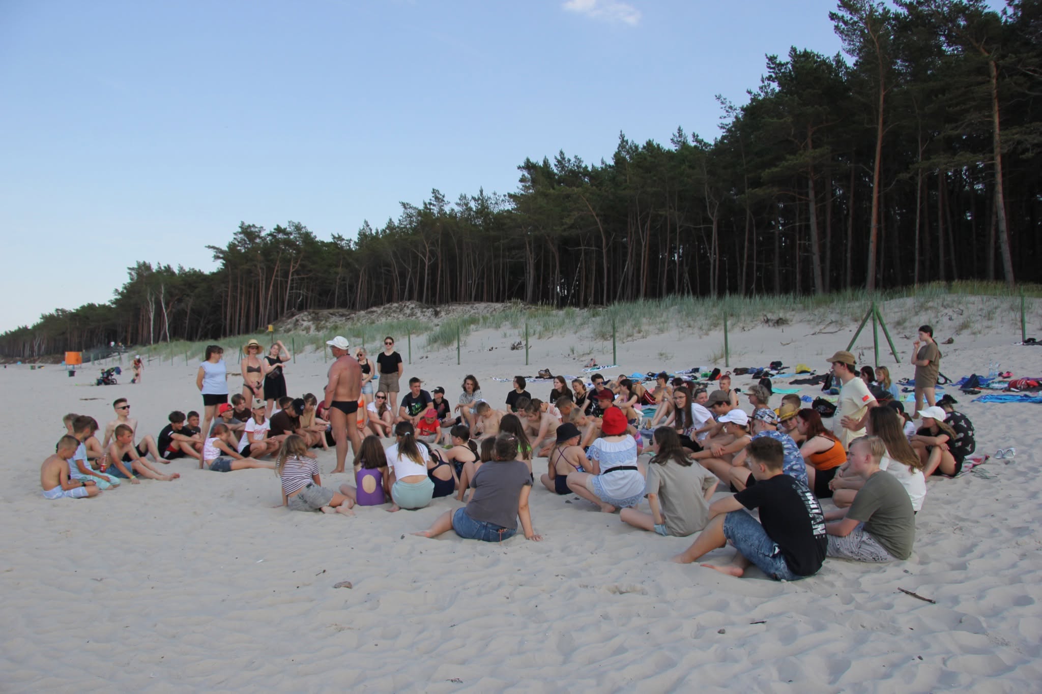 Uczestnicy siedzący w kręgu na plaży podczas zajęć grupowych na tle wydm i lasu